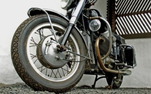 Vue en contre-plongée d'une moto vintage avec une grande roue avant, des rayons en métal et des pièces de moteur visibles, garée sur du gravier près d'un mur blanc et d'une structure en lattes de bois au Québec, idéale pour présenter les options Assurance moto.