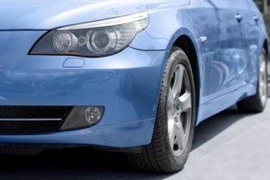 Gros plan de l'avant d'une voiture bleue, montrant le phare gauche, la roue avant et une partie du pare-chocs, garée sur une surface grise.
