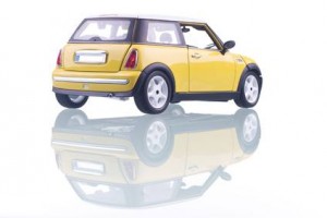 Une voiture Mini Cooper jaune vue de l'arrière gauche, avec son reflet visible sur une surface blanche brillante.