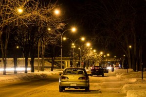 Une rue enneigée de Belœil, la nuit, est illuminée par une rangée de lampadaires. Des voitures garées, protégées par des assurances auto, bordent la route, tandis que des arbres dénudés encadrent le paysage sous la lueur jaune des lumières.