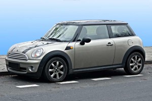 Une Mini Cooper argentée avec des garnitures noires est garée sur le bord d'une rue de Blainville, à côté d'un trottoir, sur un fond dégradé bleu, parfait pour une publicité automobile d'Assurance.