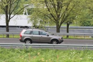 Un SUV gris roule sur une route bordée de glissières de sécurité près de Chambly, avec des arbres verdoyants et une pelouse en arrière-plan. Un camion blanc est visible derrière les arbres sur une route parallèle : une tranquillité d'esprit idéale pour votre assurance auto.