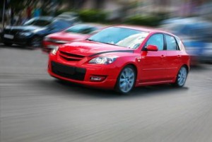 Une voiture rouge vif roule à vive allure dans une rue, l'arrière-plan étant flou pour montrer le mouvement. D'autres voitures et de la verdure sont visibles en arrière-plan.
