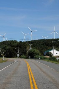 Une route rurale à double ligne jaune serpente à travers le paysage gaspésien, avec plusieurs éoliennes sur une colline verdoyante sous un ciel bleu. On aperçoit également des poteaux électriques et un bâtiment blanc, parfaits pour une photo de voiture rassurante.