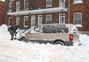 Une personne pellette la neige autour d'une fourgonnette beige garée dans une rue, enfouie sous une épaisse couche de neige. Des immeubles en briques sont visibles en arrière-plan.