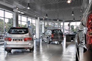 Une salle d'exposition automobile moderne avec de grandes fenêtres présente trois voitures sous des plafonniers lumineux et des sols carrelés polis, offrant un aperçu des arbres et des bâtiments - parfait pour explorer les options d'assurance automobile ou en apprendre davantage sur l'économie d'assurance.