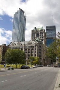 Une large rue passante, voitures et motos, passe devant un vieux bâtiment en pierre au toit végétalisé, entouré d'arbres. Derrière lui, des gratte-ciel modernes en verre s'élèvent à Montréal-Nord, sous un ciel partiellement nuageux. On aperçoit les panneaux d'Assurance auto à proximité.