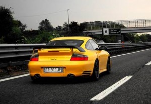 Une voiture de sport Porsche GT2 jaune vif roule sur une autoroute avec une glissière de sécurité en métal et des arbres sur le côté, s'approchant d'un viaduc avec des panneaux de signalisation visibles en arrière-plan.