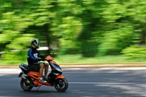 Une personne portant un casque et des vêtements décontractés conduit un scooter orange sur une route, avec des arbres verts flous en arrière-plan, suggérant un mouvement - profitant de la balade avec assurance moto pas cher pour la tranquillité d'esprit.