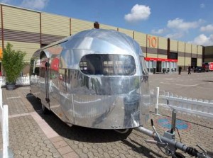 Une caravane argentée brillante aux bords arrondis est garée sur une zone pavée près d'un bâtiment moderne aux murs rayés et aux grandes fenêtres sous un ciel bleu avec des nuages.