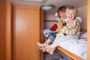 Deux jeunes enfants sont assis et sourient sur la couchette supérieure d'un lit superposé en bois, tenant des peluches dans leurs bras. La pièce est décorée de murs et de meubles en bois, avec une porte entrouverte à côté d'eux.