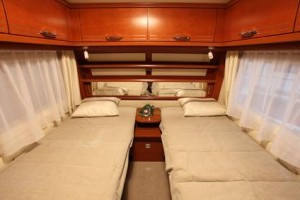 Intérieur d'un camping-car avec deux lits simples séparés par une petite table de chevet sur laquelle est posé un téléphone à cadran vert, des armoires en bois au-dessus et de grands miroirs sur le mur du fond. Des rideaux crème pendent des deux côtés des fenêtres.