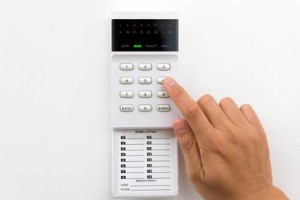 Une main appuyant sur un bouton d'un clavier d'alarme de sécurité blanc - idéal pour l'assurance habitation pas cher - monté sur un mur, doté d'un petit écran d'affichage et d'un panneau de liste de zones déroulant en dessous.