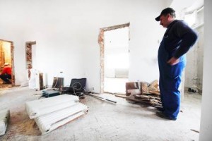 Un homme en salopette bleue se tient dans une pièce en rénovation, entouré de matériaux et d'équipements de construction. Un ouvrier est visible à travers une porte. Les murs inachevés soulignent l'importance d'une habitation sécurisée pour la protection, même pendant les travaux.