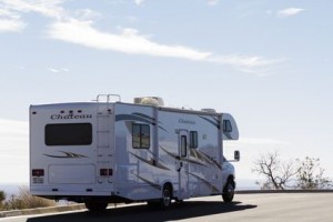 Un camping-car blanc, portant l'inscription « Château », est garé sur une route déserte, avec un ciel dégagé et l'océan au loin en arrière-plan. Une végétation clairsemée est visible le long de la route.