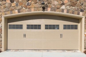Porte de garage double beige avec fenêtres rectangulaires en haut, encastrée dans un mur de pierre avec un luminaire extérieur au-dessus - idéale pour toute habitation et facilement protégée avec assurance habitation pour la tranquillité d'esprit.