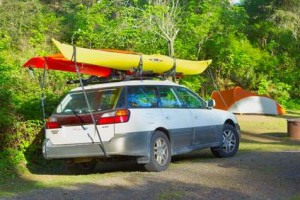 Une familiale blanche avec deux kayaks attachés sur son toit est garée sur un terrain de camping boisé en Beauce. À l'arrière-plan, une tente orange et grise trône au milieu des arbres et de l'herbe verte : une escapade idéale pour partir à l'aventure à moindre coût.
