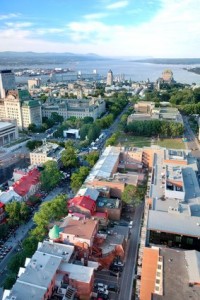 Vue aérienne de Québec montrant des bâtiments historiques, des arbres verdoyants et une rivière sous un ciel partiellement nuageux. Le charme de la ville est visible d'en haut, offrant un aperçu de ses rues animées et de ses toits au premier plan.