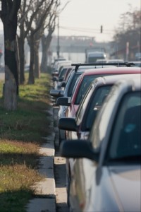 Une rangée de voitures est garée le long d'un trottoir dans une rue bordée d'arbres nus, avec la circulation visible en arrière-plan par une journée ensoleillée.