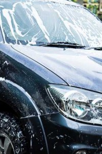 Une voiture de couleur foncée, recouverte de mousse blanche, est en cours de lavage. Le pare-brise, le capot et les phares sont visiblement savonneux, indiquant que le nettoyage est en cours.