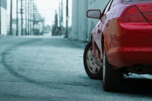 Gros plan de l'arrière d'une voiture rouge garée dans une rue de la ville avec des traces de pneus visibles sur le trottoir et des lignes électriques en arrière-plan.