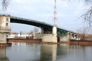 À Sorel, un pont vert et béton enjambe une rivière calme. Des bâtiments et des arbres nus sont visibles en arrière-plan sous un ciel nuageux. Une haute tour métallique rouge et blanche se dresse sur le pont, offrant une vue panoramique près des bureaux d'Assurance Véhicule Sorel.