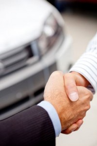 Deux personnes se serrent la main devant une voiture blanche floue, suggérant une transaction ou un accord réussi, peut-être lié à l'achat ou à la vente d'un véhicule avec des options d'assurance automobile à Anjou.