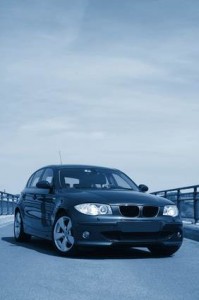 Une berline BMW de couleur foncée est garée sur une route pavée sous un ciel partiellement nuageux à Mirabel, avec des garde-corps métalliques de chaque côté. L'image est teintée de bleu, soulignant le style élégant que vous souhaitez protéger avec une voiture de qualité.