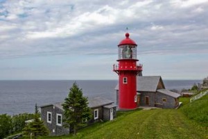 Un phare rouge vif se dresse sur une colline herbeuse à Gaspé, surplombant l'océan et les bâtiments en bois sous un ciel partiellement nuageux.