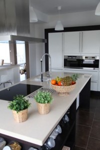 Cuisine moderne avec îlot central blanc, panier de fruits frais, deux herbes aromatiques en pot et électroménagers intégrés. L'espace est épuré et lumineux grâce à ses suspensions et à son design contemporain.