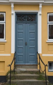 Porte en bois bleu surmontée de ferronnerie décorative, située dans un bâtiment peint en jaune et orné de moulures blanches. L'entrée est dotée de quelques marches en pierre et de rampes en métal noir de chaque côté. Deux fenêtres encadrent la porte.