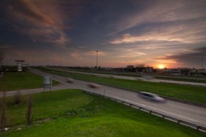 Une autoroute près de Montréal au coucher du soleil, avec des voitures floues qui filent à toute vitesse, de l'herbe verte de chaque côté et un ciel coloré parsemé de nuages. Des bâtiments et quelques panneaux – peut-être pour « assurance auto moins cher » – sont visibles au loin.
