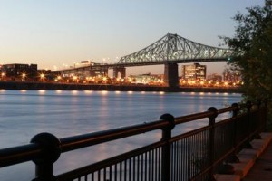 Un pont en acier illuminé au crépuscule enjambe une rivière calme à Montréal, avec des bâtiments de la ville et des lampadaires brillants en arrière-plan, et une clôture en métal noir au premier plan, capturant le charme de la ville où la vie peut être moins chère.