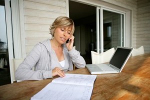Une femme est assise à une table en bois avec un ordinateur portable et un cahier ouverts, parlant sur son téléphone portable, comparant peut-être les options d'assurance habitation moins chère tout en travaillant ou en étudiant dans son intérieur de maison moderne.