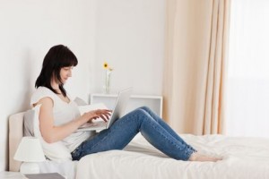 Une femme est assise sur un lit, les jambes allongées, utilisant un ordinateur portable, comparant peut-être les options d'assurance habitation moins chère. La pièce est lumineuse et minimaliste, avec une table de chevet blanche, une lampe et un vase avec des fleurs jaunes en arrière-plan.