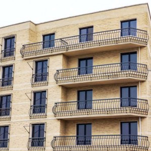 Un immeuble d'appartements moderne avec des murs en briques jaunes et des garde-corps en métal noir sur plusieurs balcons, doté de grandes portes vitrées à chaque étage, géré par un syndicat pour la copropriété.
